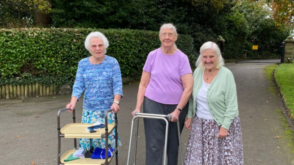 Three older people walking, two with teh help of walking frames